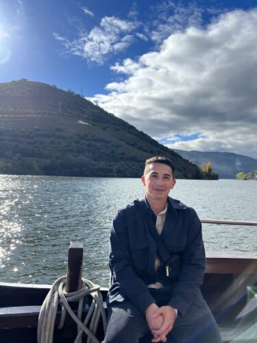 A person sits on a boat with a lake and mountains in the background. Its a sunny day with clouds in the sky. The person is smiling and wearing a dark jacket.